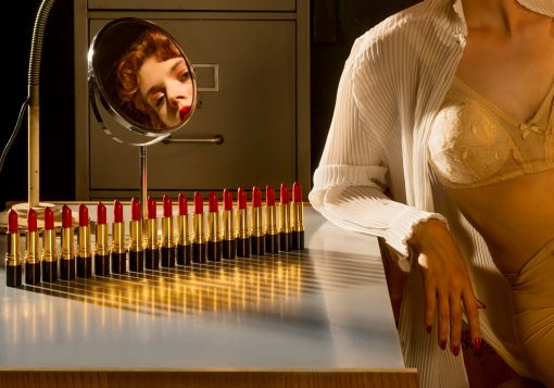 Für dieser Fotografie dient eine Reihe Lippenstifte als Symbol für Munition. Eine laszive Frau sitzt in Unterwäsche gelangweilt an einem Tisch und hält eine Pistole in der Hand.