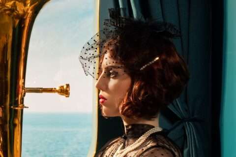 Horst Kistner La Traversata photography Detail 1 Zeigt das Porträt einer Jungen Musikerin. Sie spielt Tuba und halt ihr Instrument und sitzt an einem Fenster eines Kreuzfahrtschiffes. Draußen sieht man das blaue Meer.