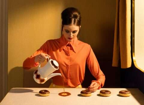 Horst Kistner The Student zeigt eine Frau die Kaffee auf einen Tisch verschüttet. Sie scheint es jedoch mit Absicht zu tun. Schokodonuts liegen in einer Reihe. Diese inszenierte Fotografie ist ei surreales Meisterstück.