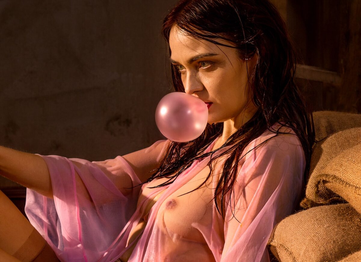 Das Detail der Fotografie von Horst Kistner After The Flood zeigt eine Frau mit verschleierten, nackten Brüsten. Sie lehnt an Sandsäcken und macht mit pinkfarbenen Kaugummi eine Blase. Ihre Haare und der Körper sind nass.