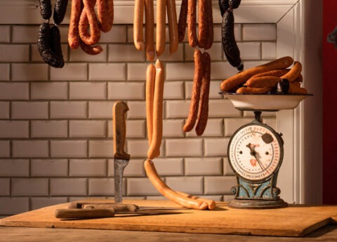 Detailaufnahme aus Horst Kistners Fotografie Delicatessen mit Küchenwaage, Messer, aufgehängten Würsten und weiß gekachelter Wand.