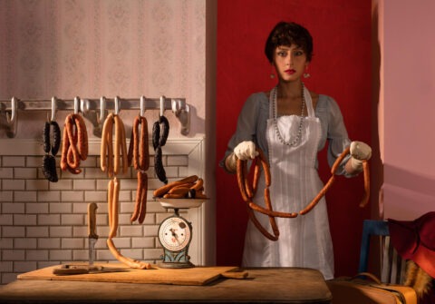 Horst Kistner Delicatessen im Vollformat ist eine Fotografie mit Frau in weißer Schürze vor roter Wand, aufgehängten Würsten, Messer und alter Küchenwaage in surrealer Küchenszene.