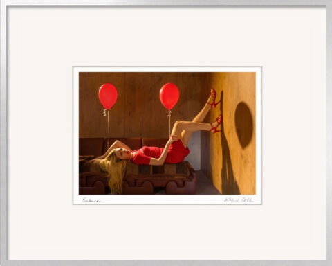 Fotografie „Balance“ von Horst Kistner mit einer Frau im roten Kleid auf einem Sofa, gehalten von zwei roten Ballons. Die Szene vermittelt Leichtigkeit, Gleichgewicht und Freiheit. Präsentiert im silbernen Aluminiumrahmen.