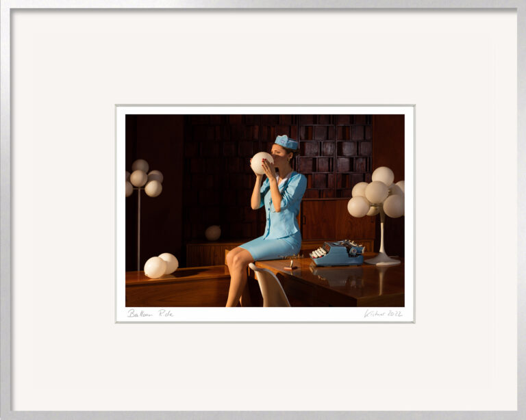 Fotografie „Balloon Ride“ von Horst Kistner mit einer Frau in hellblauer Uniform, die einen weißen Ballon aufbläst. Umgeben von weiteren Ballons und einer Schreibmaschine entsteht eine poetische Szene zwischen Büroalltag und Fantasie. Präsentiert im silbernen Aluminiumrahmen.